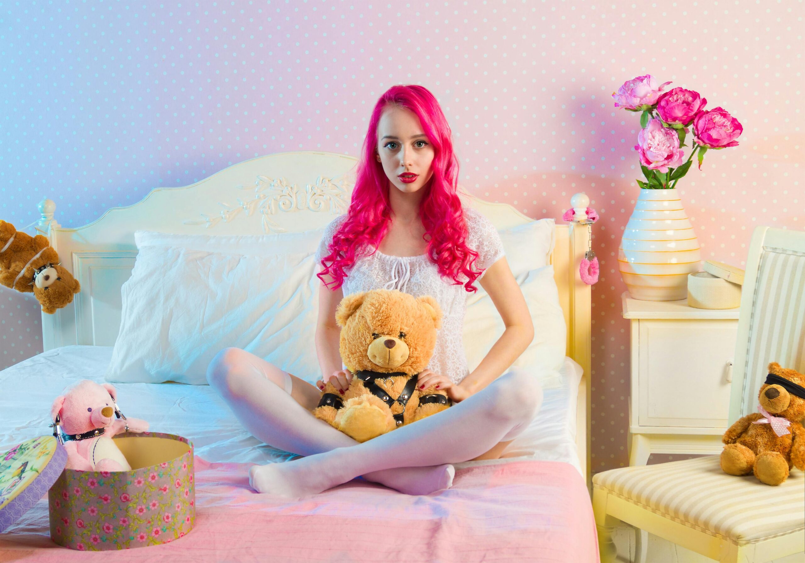 Stylized indoor portrait of a pink-haired woman sitting with teddy bears in a bright, whimsical bedroom.