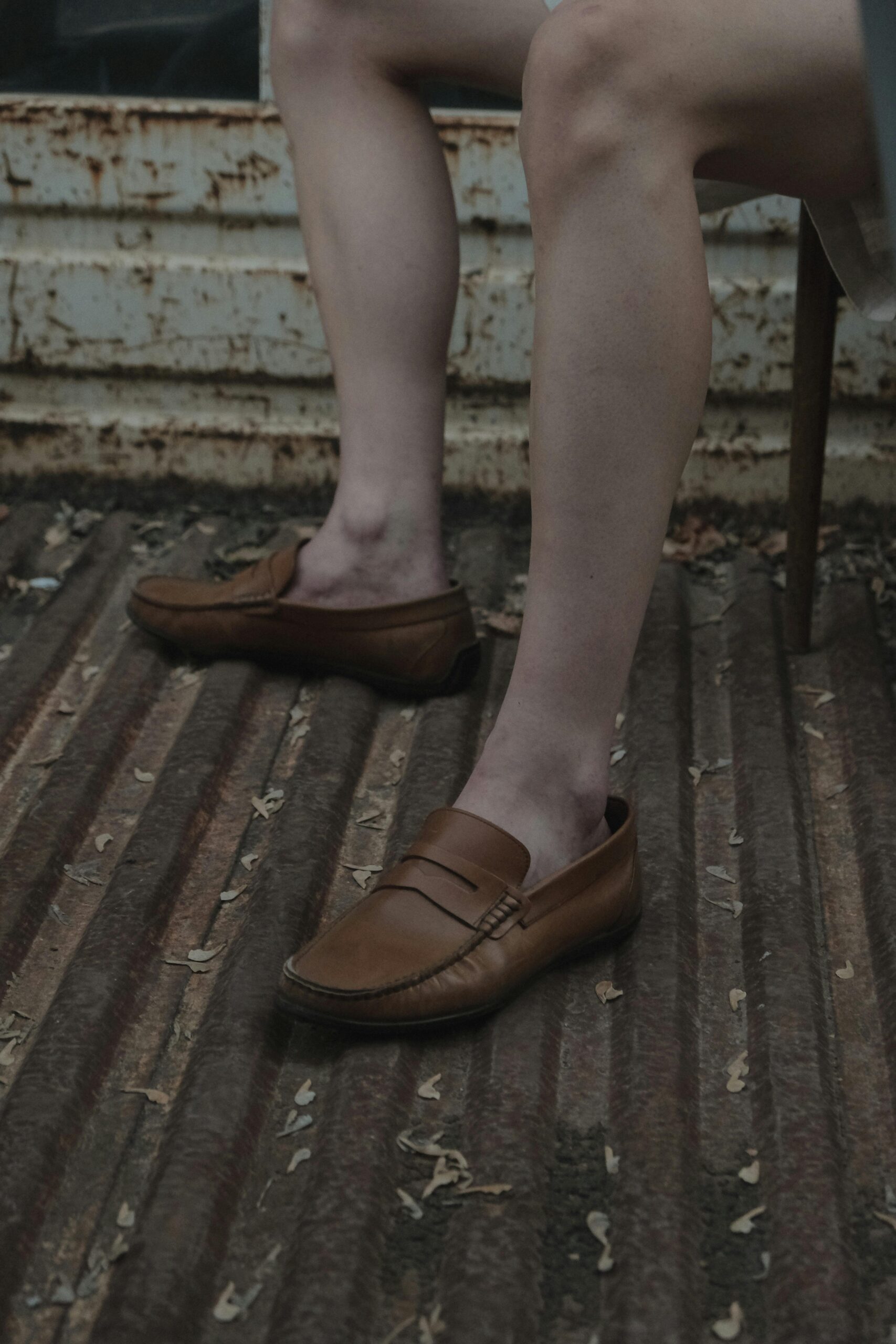 Close-up of stylish brown leather loafers worn by person sitting on metal surface with rustic backdrop.