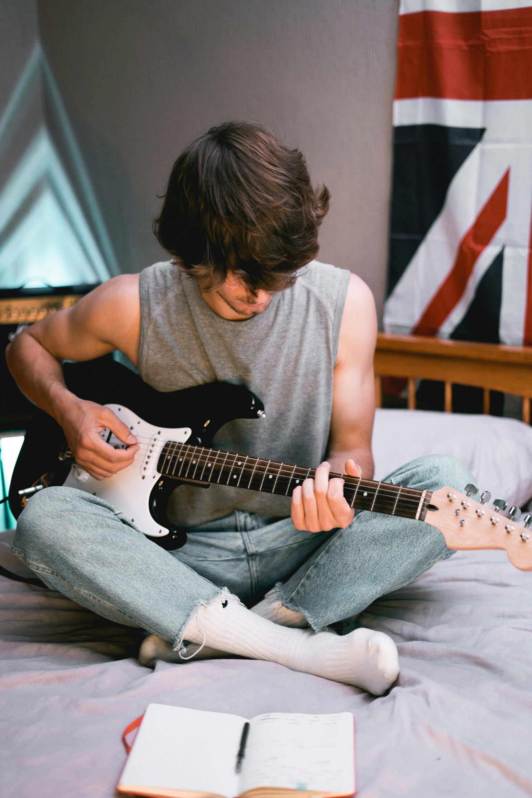 Chilled young adult playing electric guitar on bed, notebook open, Union Jack in background.