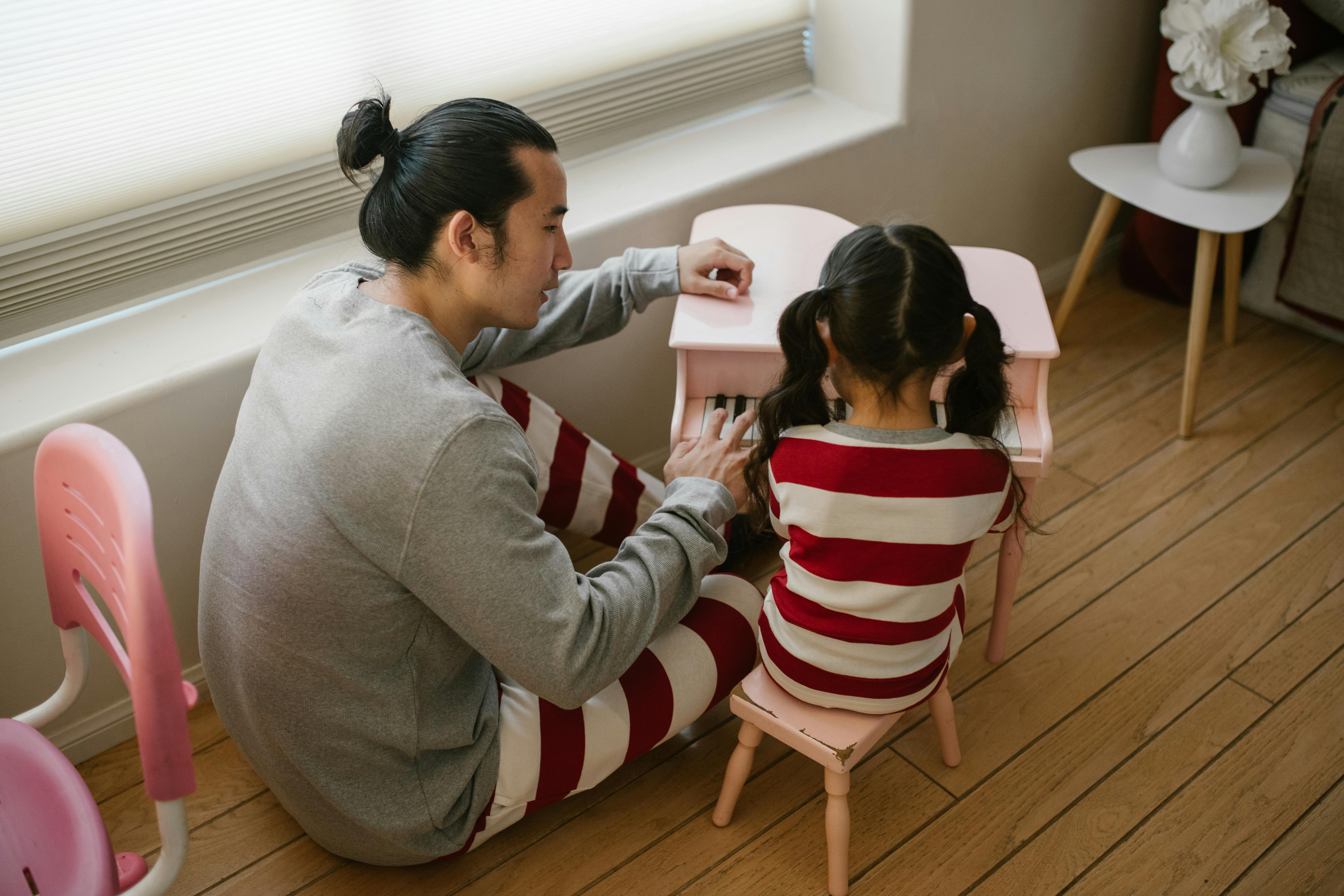 A father teaches his young daughter to play a small pink piano in a cozy room.