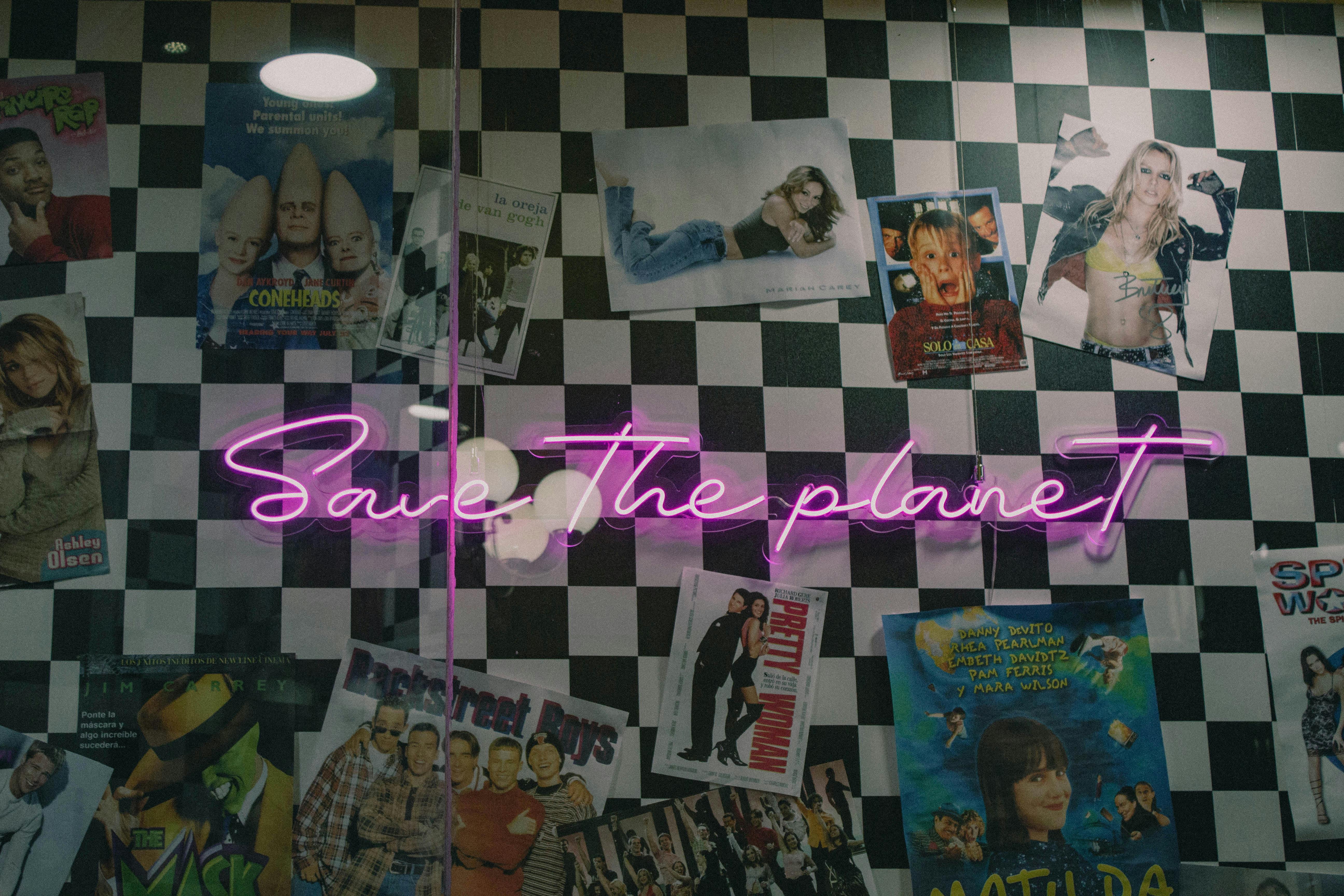 Neon sign with 'Save The Planet' on a checkerboard wall, surrounded by vintage posters.