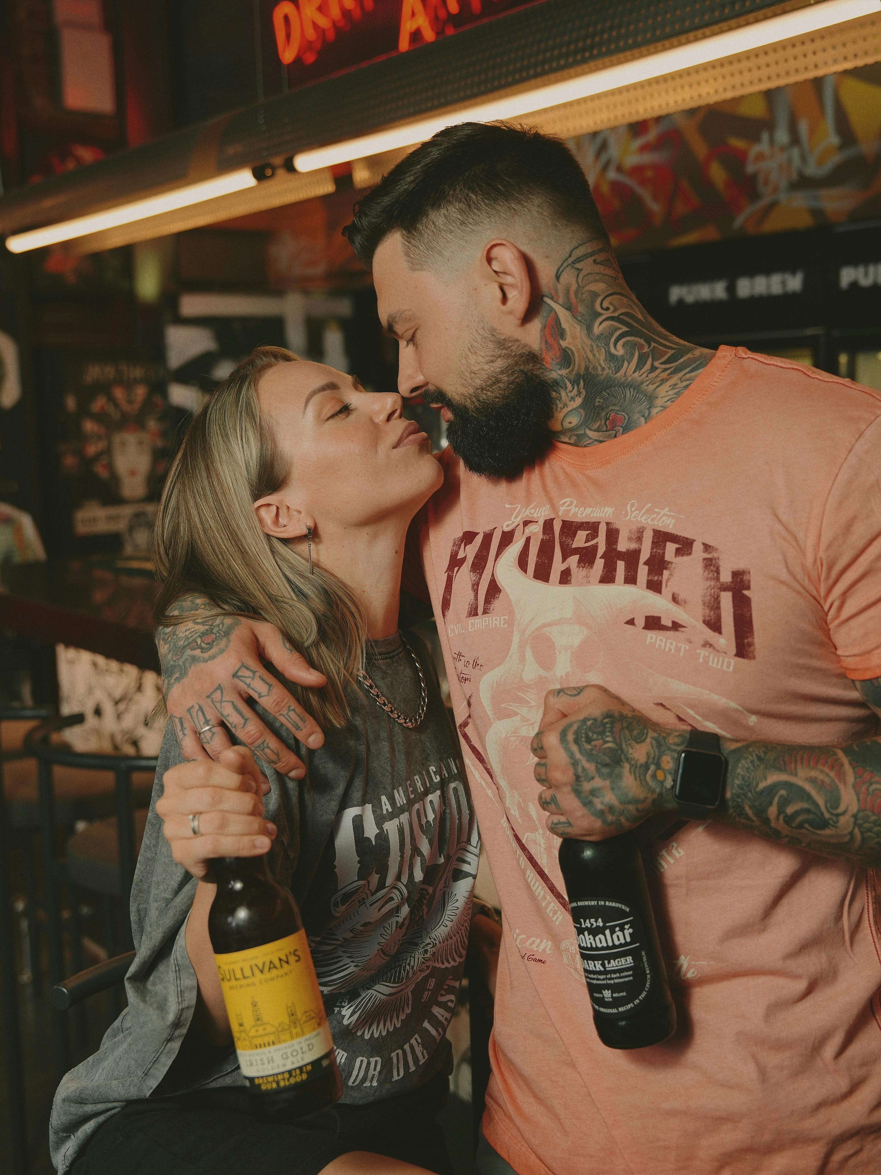 A young, tattooed couple embraces while holding beer bottles at a bar.