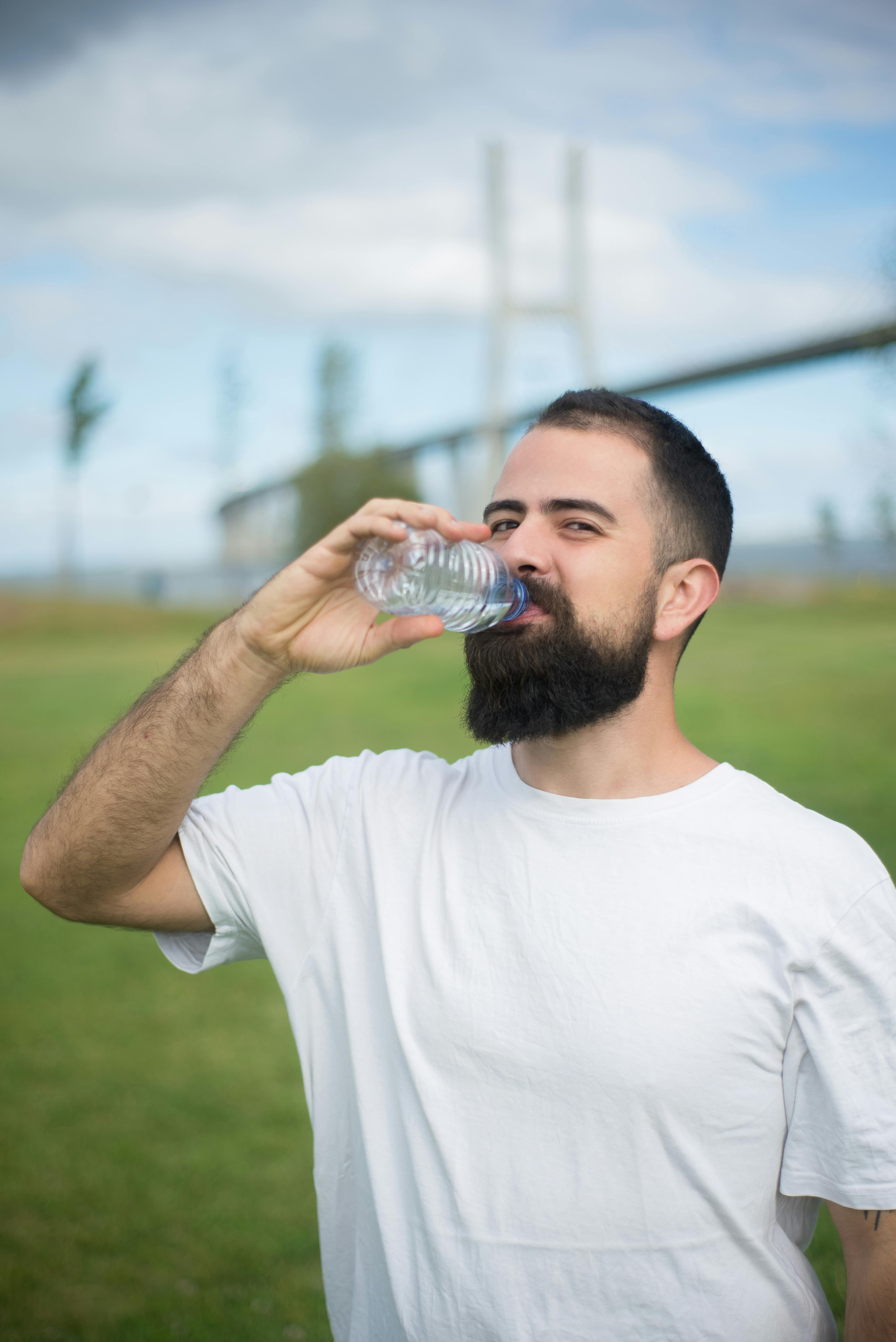 Portrait of a bearded man drinking water outdoors on a sunny day, bridge in the background.